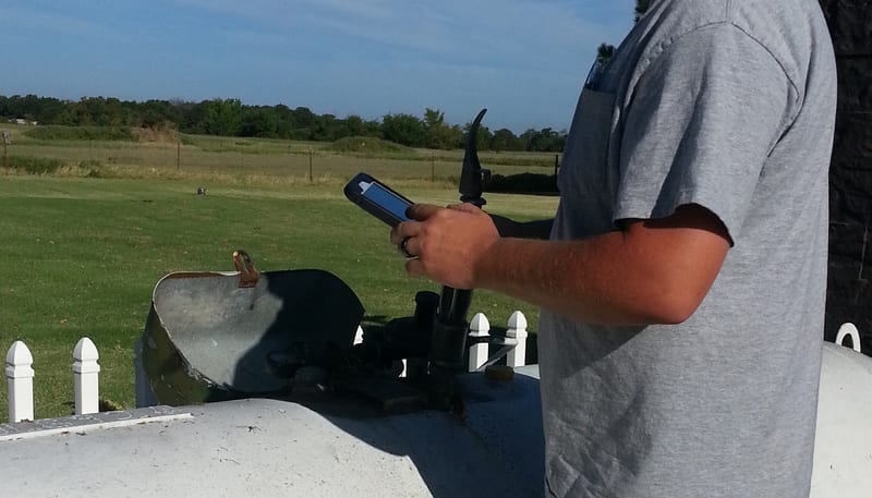 A propane technician's hand holds a rugged handheld device beside the open service dome of a residential propane tank, verifying tank data in the field.