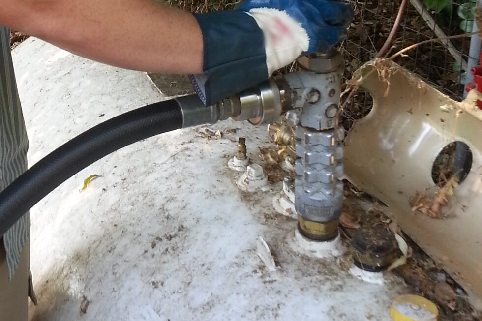 A propane delivery hose coupled to the fill valve on a weathered residential ASME tank, a gloved hand steadying the connection.